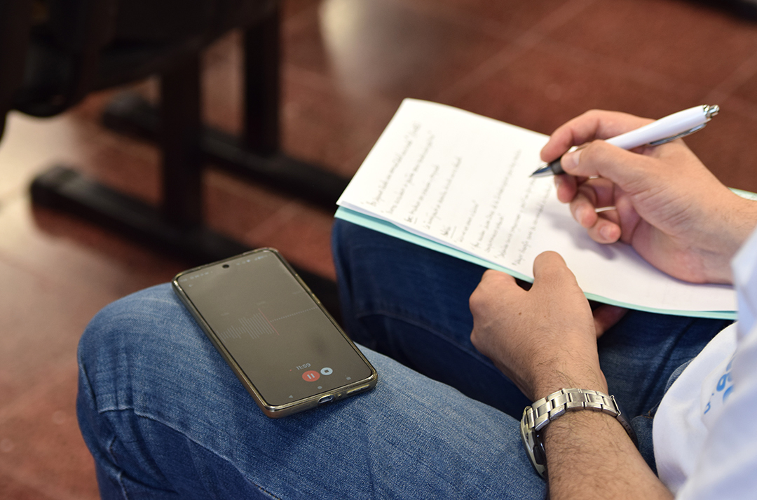 mano-de-una-persona-escribiendo-sobre-un-papel-y-con-un-celular-reproduciendo-un-audio