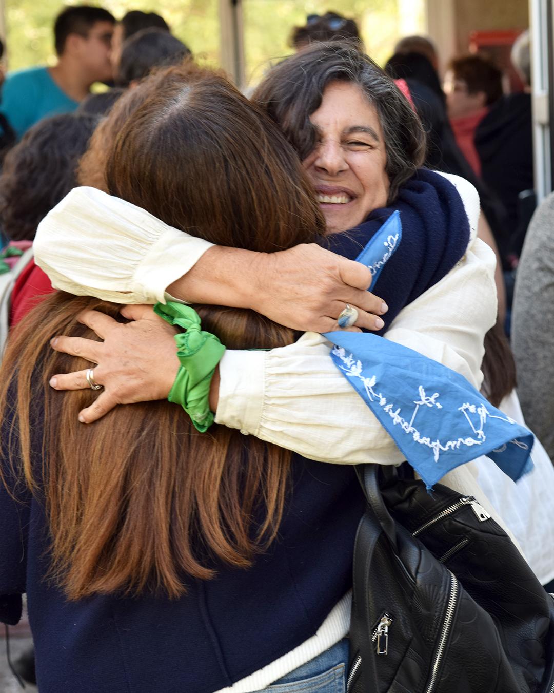 En primer plano, abrazo entre dos personas, se ve la cara de la decana entrante con una sonrisa y los pañuelos azul en la mano derecha y un pañuelo verde en la muañeca de mano izquierda