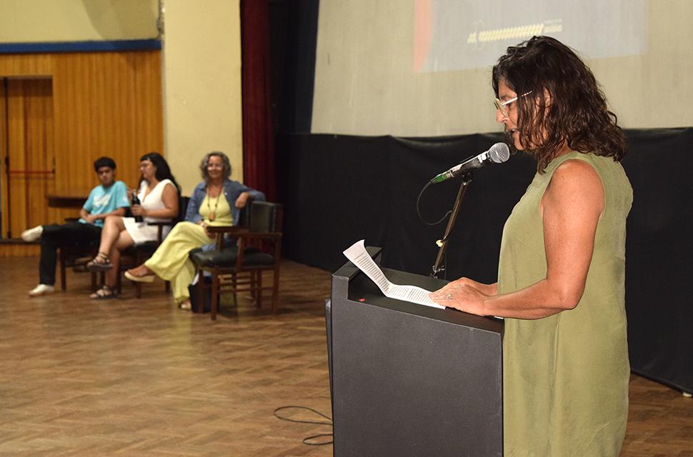 En primer plano, de perfil  se encuentra la Decana Aljandra Dominguez, leyendo su discurso de bienvenida, de fondo se ven a la secretaria académica, secrretaria de asuntos estudiantiles y presidente de centro de estudiantes, sentados con la mirada dirigida hacia la decana