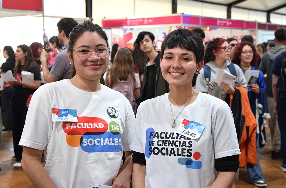 Dos estudiantes de Sociales en primer plano, frente a cámara sonriendo, en el fondo hay personas caminando