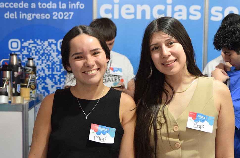 Dos estudiantes de facultad de sociales frente a cámara sonriendo, en el stand